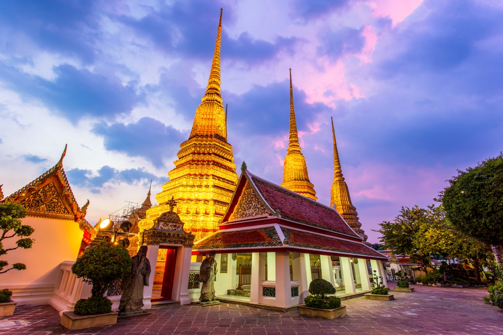 ยอดแหลมสีทองของวัดโพธิ์หรือวัดพระนอนใน วัดที่สวยที่สุดในประเทศไทย กรุงเทพฯ เรืองแสงในยามพลบค่ำตัดกับท้องฟ้าสีม่วงอมชมพู ล้อมรอบด้วยต้นไม้อันเงียบสงบ.