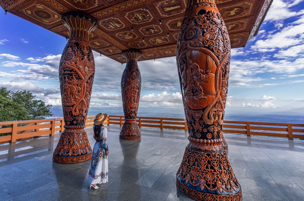 ผู้หญิงสวมชุดและหมวกยืนอยู่ใต้ ภูเขาสูงสุดในไทย ศาลาไม้แกะสลักที่มีเสาประดับอย่างวิจิตร มองออกไปเห็นท้องฟ้าที่มีเมฆครึ้มและเส้นขอบฟ้า ชวนให้นึกถึงทิวทัศน์จากพระอุโบสถสูงสุดในไทย