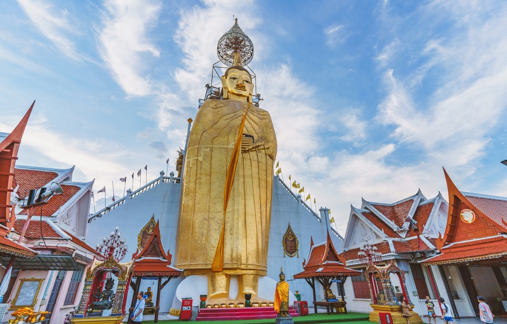 พระพุทธรูปสีทององค์ใหญ่ตั้ง รอยพระพุทธบาท ตระหง่านสง่างามอยู่หน้าวิหาร ท่ามกลางความสงบนิ่ง สะท้อนถึงความสำคัญทางจิตวิญญาณของรอยพระพุทธบาท ท่ามกลางท้องฟ้าสีฟ้าสดใสและเมฆที่กระจัดกระจาย พระพุทธรูปองค์นี้เชื้อเชิญให้ผู้มาเยี่ยมชมสำรวจรอยพระพุทธบาทอันศักดิ์สิทธิ์ในบริเวณใกล้เคียง