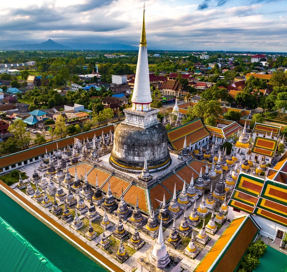 มุมมองทางอากาศ วัดที่สวยที่สุดในประเทศไทย ของเจดีย์พุทธขนาดใหญ่ล้อมรอบด้วยเจดีย์ขนาดเล็กในกลุ่มวัดที่วัดพระนอน ตั้งอยู่ในตัวเมืองที่มีต้นไม้เขียวขจีและภูเขาไกลๆ ภายใต้ท้องฟ้าที่มีเมฆบางส่วน.