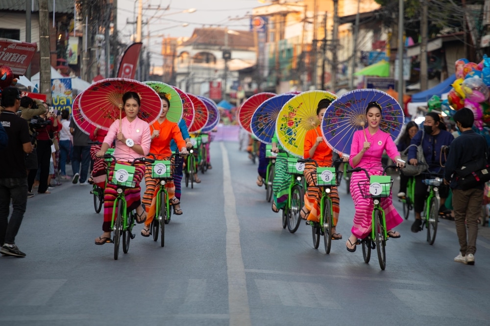 กลุ่มคนสวมชุดไทยสีสันสวยงาม ขี่จักรยานที่ประดับด้วยร่มกระดาษสีสดใส เฉลิมฉลองในขบวนแห่ริมถนนเพื่อเชิดชูความงามอันยิ่งใหญ่ตระการตาของสิ่งศักดิ์สิทธิ์สูงสุดในไทย.