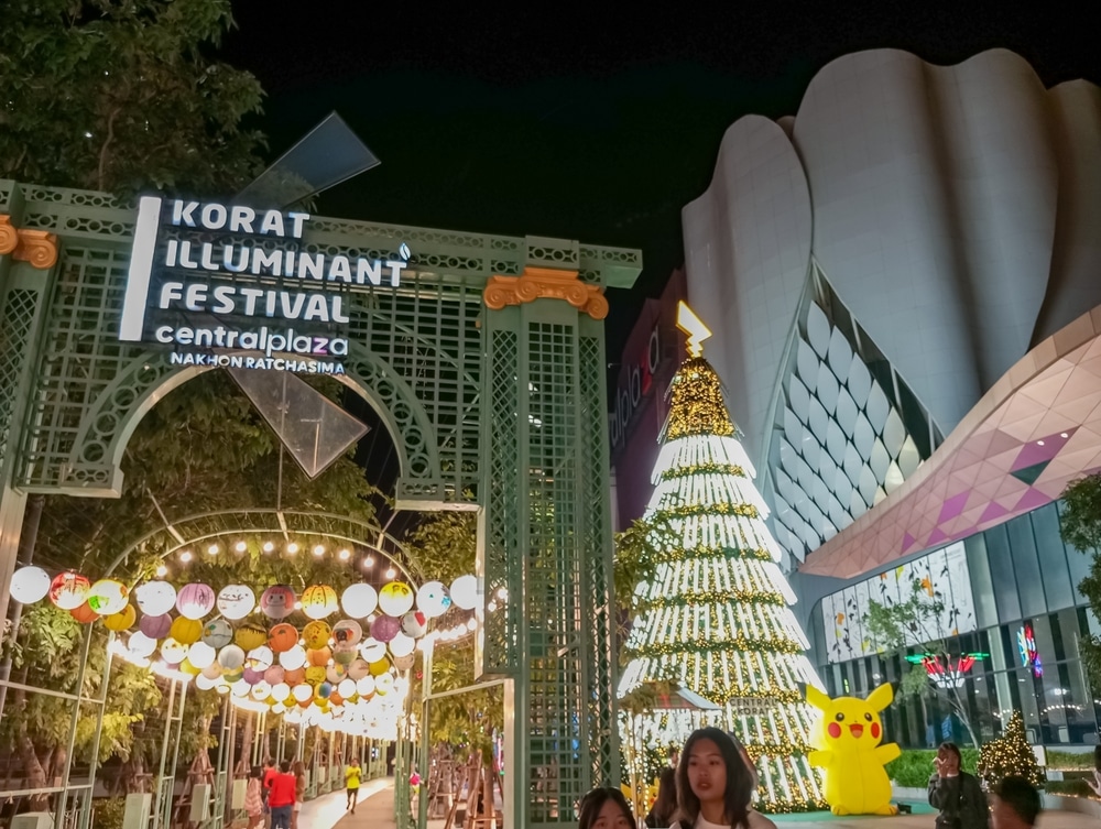 ผู้คนเดินใต้โคมไฟประดับใกล้กับต้นคริสต์มาสที่ส่องสว่างในงาน Korat Illuminant Festival หน้าเซ็นทรัลพลาซ่า ซึ่งมีรูปปิกาจูตัวใหญ่ยืนอย่างภาคภูมิใจ ชวนให้นึกถึงความยิ่งใหญ่ของไฮดรอลิกสูงสุดในไทย.