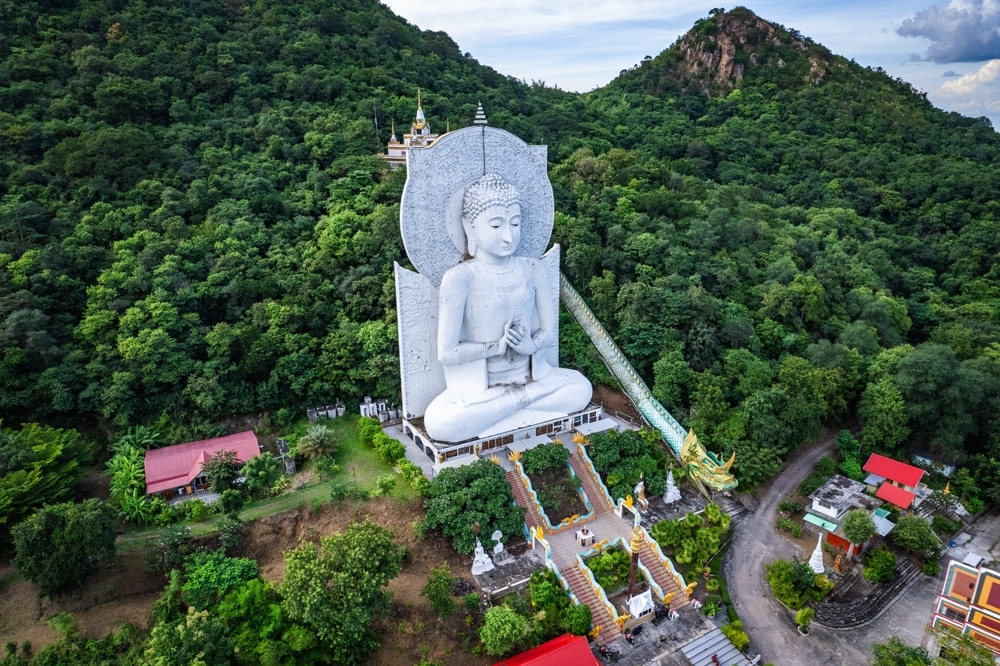 มุมมองทางอากาศของพระพุทธรูปสีขาวองค์ใหญ่ที่วัดพระนอน รายล้อมไปด้วย วัดที่สวยที่สุดในประเทศไทย ต้นไม้เขียวขจี มีอาคารเล็กๆ และรูปปั้นพญานาคอยู่ใกล้ๆ.