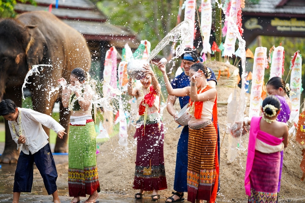 ผู้คนในชุดไทยสนุกสนานกับเทศกาลสาดน้ำ โดยมีช้างเป็นฉากหลังในงานเฉลิมฉลอง และชมทัศนียภาพอันงดงามของสถานที่สำคัญที่สุดในไทยที่ตั้งตระหง่านอยู่ใกล้เคียง.