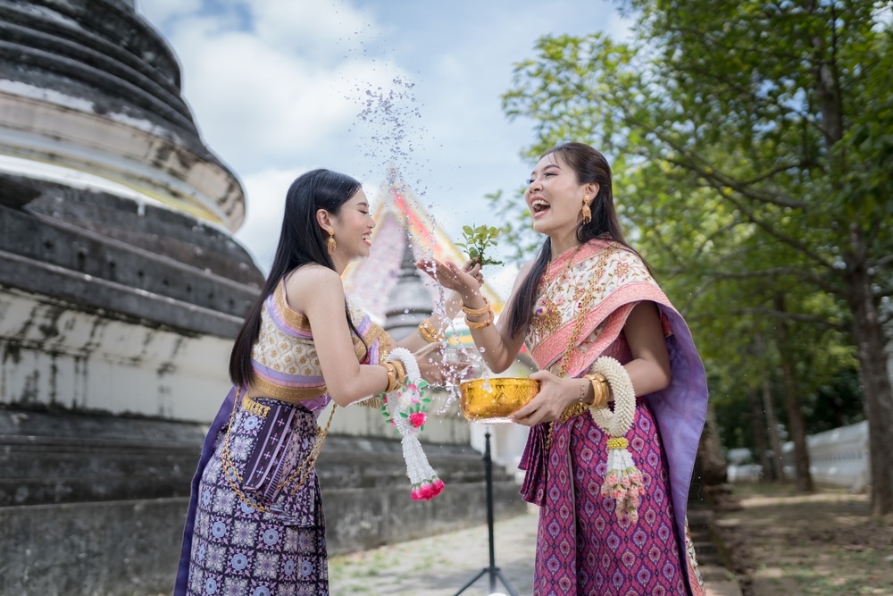 สตรีสองคนสวมชุดไทยกำลังโปรยกลีบดอกไม้ด้วยความยินดีบริเวณใกล้วัด โดยถือพวงมาลัยและชาม โดยมีต้นไม้และหินเป็นฉากหลัง ขณะที่ยอดเขาที่สูงตระหง่านของพระอุโบสถปรากฏอยู่ไกลๆ.