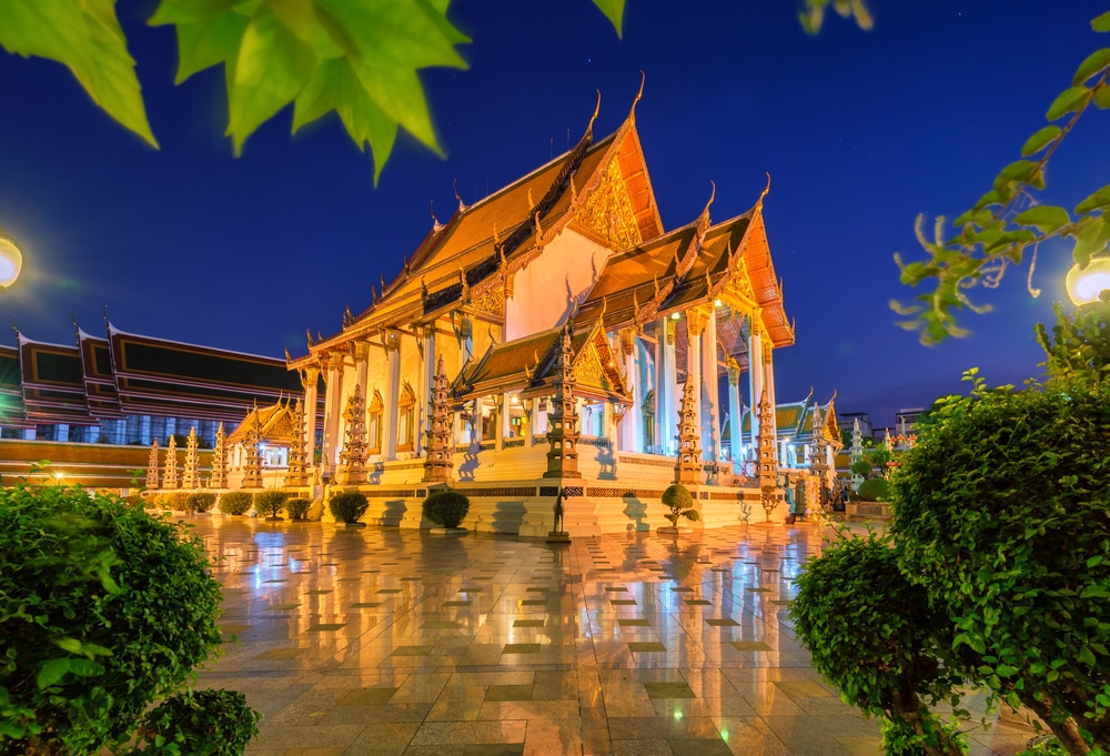 วัดไทยที่ รอยพระพุทธบาท สว่างไสวในตอนกลางคืน ล้อมรอบด้วยแมกไม้เขียวขจีและสะท้อนบนพื้นเปียกมันวาว ชวนให้นึกถึงบรรยากาศอันเงียบสงบของรอยพระพุทธบาท