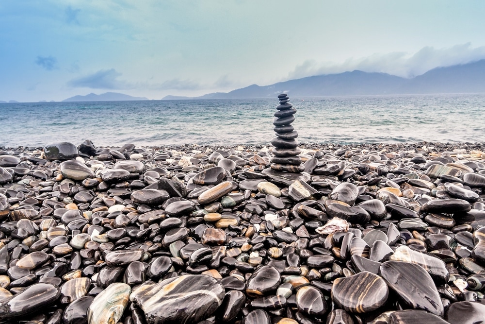 กองหินสีเข้มเรียบๆ วางอยู่บนชายหาดที่เต็มไปด้วยกรวดในภาคใต้ ภายใต้ ท่องเที่ยวภาคใต้ ท้องฟ้าที่มีเมฆครึ้มและมีภูเขาไกลๆ ข้ามทะเลไป