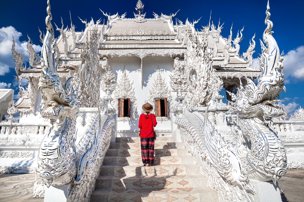 บุคคลที่สวมเสื้อสีแดงยืน วัดที่สวยที่สุดในประเทศไทย หันหน้าไปทางวัดสีขาวหรูหราที่มีการแกะสลักอย่างประณีตใต้ท้องฟ้าสีคราม เตรียมไหว้พระนอน ณ วัดพระนอนอันน่าทึ่ง.