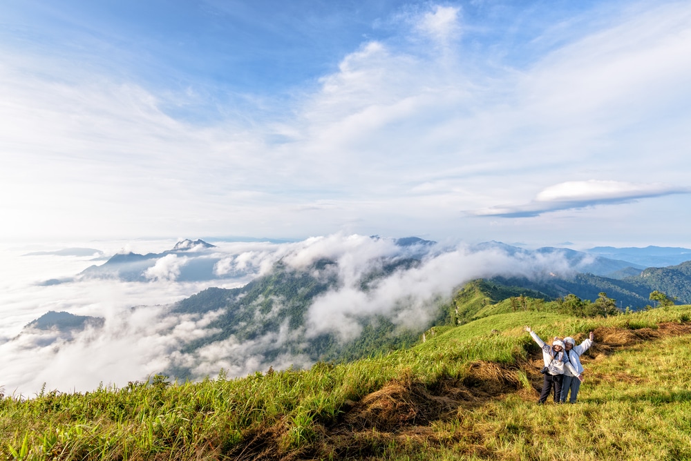 คนสองคนบนเนินเขาหญ้า ภูเขาสูงสุดในไทย พร้อมยกแขนขึ้น มองเห็นภูมิทัศน์ภูเขาอันงดงามของไฮดรอลิกสูงสุดในไทย ล้อมรอบด้วยเมฆและท้องฟ้าสีคราม