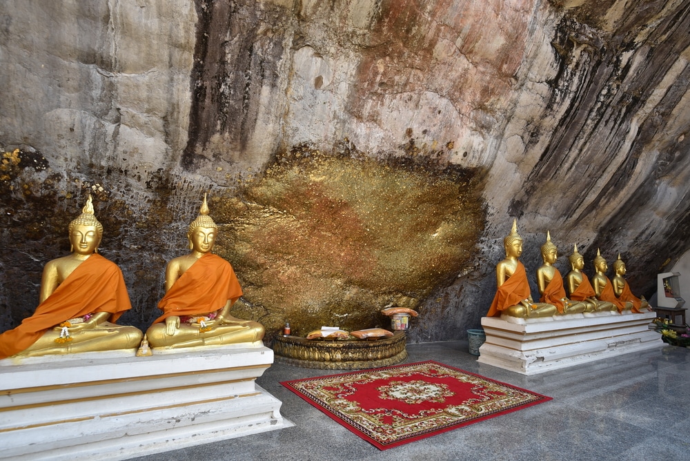 พระพุทธรูป รอยพระพุทธบาท สีทองอร่าม สวมจีวรสีส้ม ประดิษฐานอยู่ในถ้ำ ท่ามกลางแสงนวลอ่อนๆ ด้านหน้ามีพรมแดงทองวางอย่างสง่างามใต้รอยพระพุทธบาทอันศักดิ์สิทธิ์