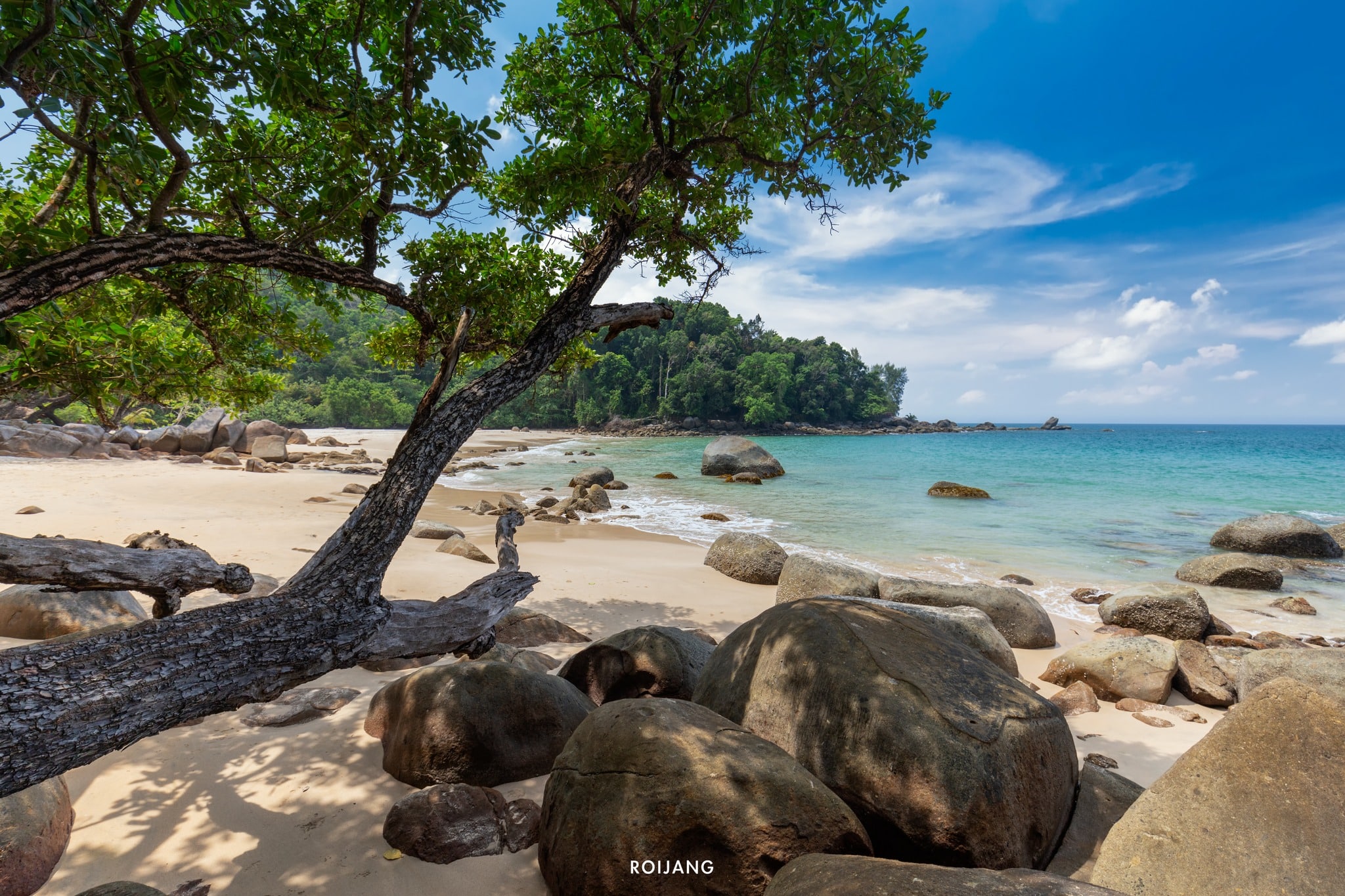 ชายหาดหินมีก้อนหินขนาดใหญ่ น้ำทะเลสีฟ้าใส และต้นไม้โค้งเหนือผืนทราย โดยมีฉากหลังเป็นเนินเขาสีเขียวขจีและท้องฟ้าสีฟ้า เหมาะสำหรับ จุดกางเต็นท์พังงา.