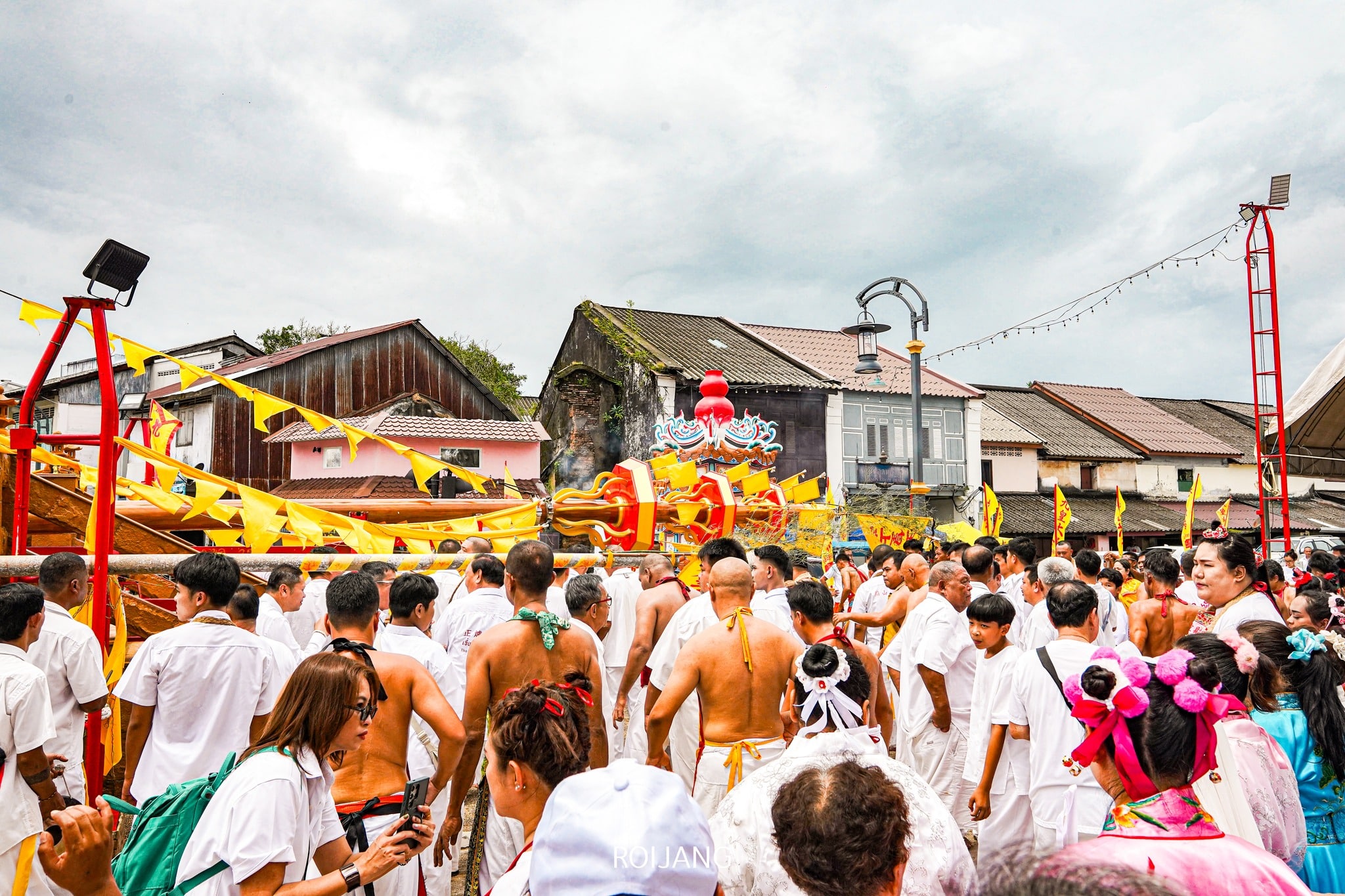 ฝูงชนสวมชุดสีขาวมารวมตัวกันรอบ ๆ ร่างใหญ่สีสันสดใสบนเรือที่ประดับด้วยธงสีเหลือง โดยมีฉากหลังเป็นบ้านเรือนและท้องฟ้าที่มีเมฆครึ้ม ทุกสิ่งทุกอย่างดำเนินไปราวกับฉากภายใต้ความยิ่งใหญ่อันไกลโพ้นของพระผู้เป็นเจ้าสูงสุดในไทย.