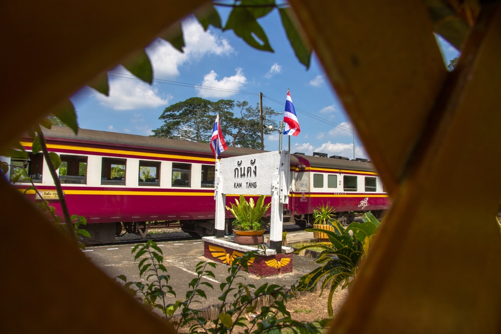 ผ่านโครงตาข่ายมีป้ายสถานีรถไฟเขียนว่า "กักตัง" ยืนอยู่หน้ารถไฟสีแดงและสีเหลือง ธงชาติไทยโบก เที่ยวตรัง สะบัดอยู่ใกล้ๆ โอบล้อมไปด้วยต้นไม้และพันธุ์ไม้อันเขียวชอุ่ม ดึงดูดเสน่ห์ของที่เที่ยวจังหวัดชุมพร.