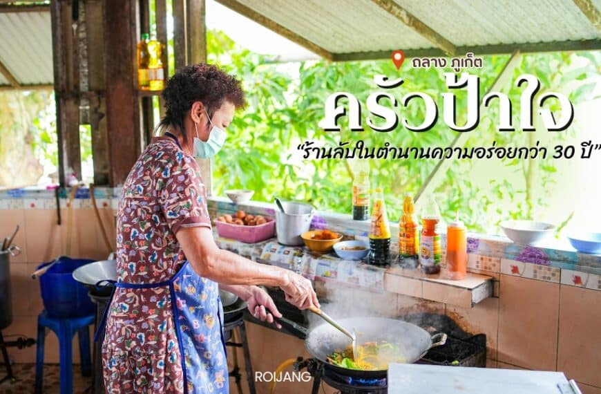 มีคนกำลังทำอาหารอยู่ที่เคาน์เตอร์ครัวที่รายล้อมไปด้วยวัตถุดิบและอุปกรณ์ต่างๆ บรรยากาศร้านชวนให้นึกถึงครัวป้าใจ ให้บรรยากาศกลางแจ้งพร้อมความเขียวขจีเป็นฉากหลัง คล้ายกับมุมเงียบสงบของร้านอาหารภูเก็ต.