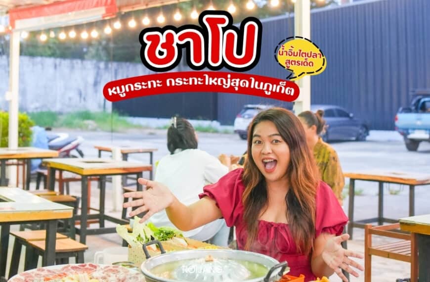 ผู้หญิงคนหนึ่งนั่งอยู่ที่โต๊ะร้านอาหารกลางแจ้งในย่านตลาดนาคาภูเก็ต รายล้อมไปด้วยอาหารหลากหลายอย่างชาโปและหมูกระทะ เธอยิ้มและทำท่าทางตื่นเต้น ขณะที่โฆษณาอาหารที่มีชีวิตชีวาแสดงอยู่ด้านหลัง.