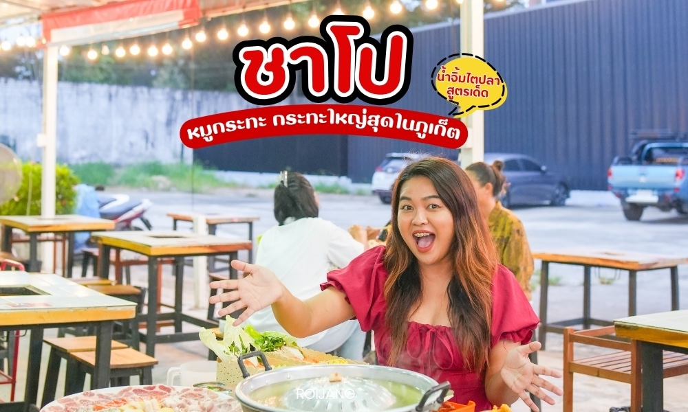 ผู้หญิงคนหนึ่งนั่งอยู่ที่โต๊ะร้านอาหารกลางแจ้งในย่านตลาดนาคาภูเก็ต รายล้อมไปด้วยอาหารหลากหลายอย่างชาโปและหมูกระทะ เธอยิ้มและทำท่าทางตื่นเต้น ขณะที่โฆษณาอาหารที่มีชีวิตชีวาแสดงอยู่ด้านหลัง.