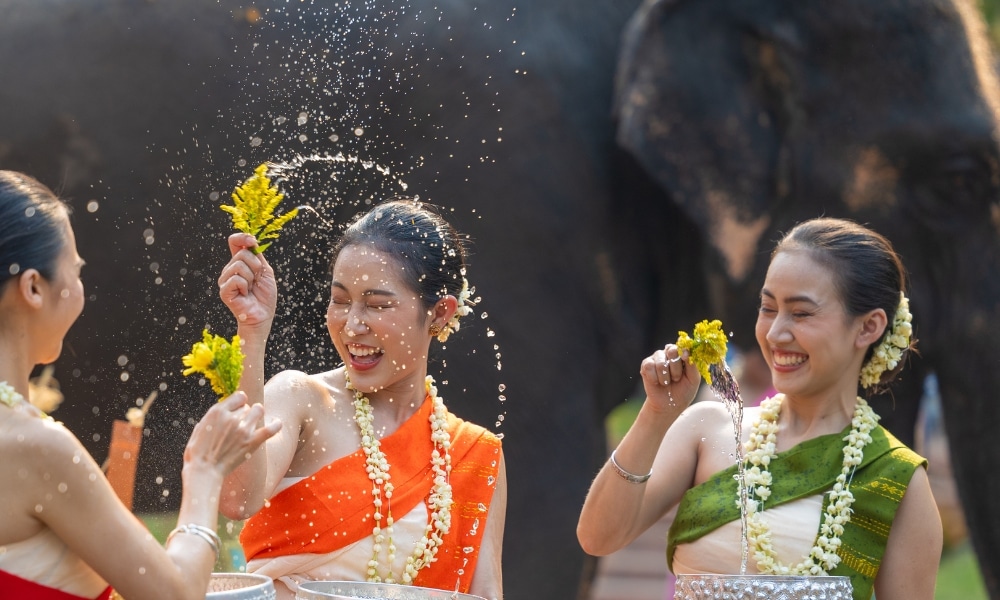 สตรีสามคนสวมชุดประจำชาติสีสันสดใสเข้าร่วมงานเทศกาลน้ำอย่างสนุกสนาน โดยถือดอกไม้ และมีช้างเล่นน้ำอย่างสนุกสนานอยู่ใกล้ๆ และมีพระพิทักษ์สูงสุดในไทยที่ตั้งตระหง่านอยู่เบื้องหลัง.