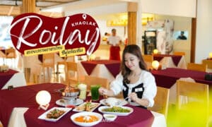 ผู้หญิงคนหนึ่งนั่งอยู่ที่โต๊ะร้านอาหาร Roilay หรอยเล พร้อมอาหารหลายอย่าง รวมทั้งซุป ผัก และเครื่องดื่ม เธอถือส้อมเตรียมจะกิน ร้านอาหารแห่งนี้มีโต๊ะไม้และตกแต่งภายในแบบสว่างสดใส.