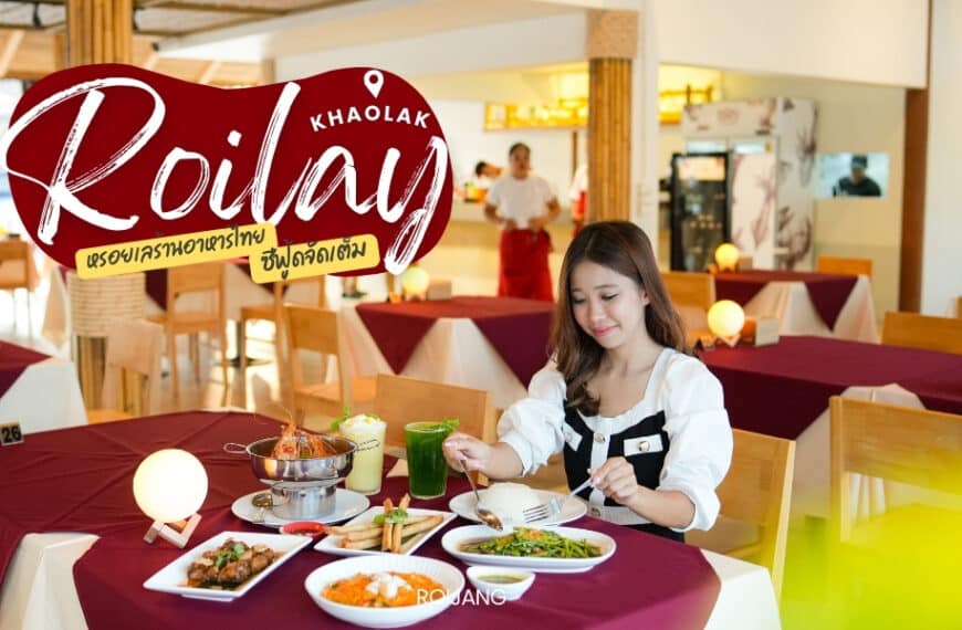 ผู้หญิงคนหนึ่งนั่งอยู่ที่โต๊ะร้านอาหาร Roilay หรอยเล พร้อมอาหารหลายอย่าง รวมทั้งซุป ผัก และเครื่องดื่ม เธอถือส้อมเตรียมจะกิน ร้านอาหารแห่งนี้มีโต๊ะไม้และตกแต่งภายในแบบสว่างสดใส.