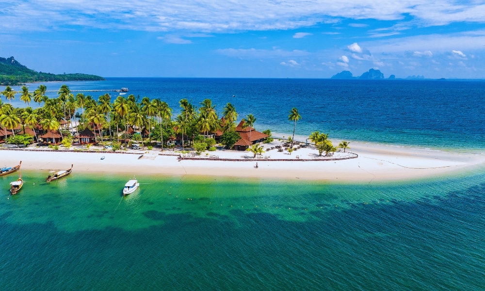 วิวทางอากาศของเกาะเขตร้อนใน ที่เที่ยวตรัง ชุมพร มีต้นปาล์ม หาดทราย และเรือหลายลำจอดเทียบท่าใกล้ชายฝั่ง มหาสมุทรสีฟ้าทอดยาวไปจนถึงเกาะห่างไกลภายใต้ท้องฟ้าที่มีเมฆบางส่วน ถือเป็นอัญมณีสำหรับผู้ที่แสวงหาความงามของสถานที่ท่องเที่ยวจังหวัดชุมพรอย่างแท้จริง.