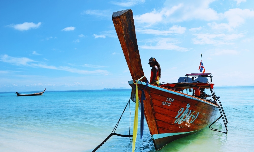 เรือไม้ที่ตกแต่งด้วยสีสันสดใสจอด ที่เที่ยวตรัง อยู่ในน้ำทะเลสีฟ้าใสในวันที่อากาศสดใส มองเห็นแหล่งท่องเที่ยวชุมพรได้อย่างสมบูรณ์แบบ เรืออีกลำมองเห็นได้แต่ไกล แสดงให้เห็นเสน่ห์ของพื้นที่ท่องเที่ยวจังหวัดชุมพร.