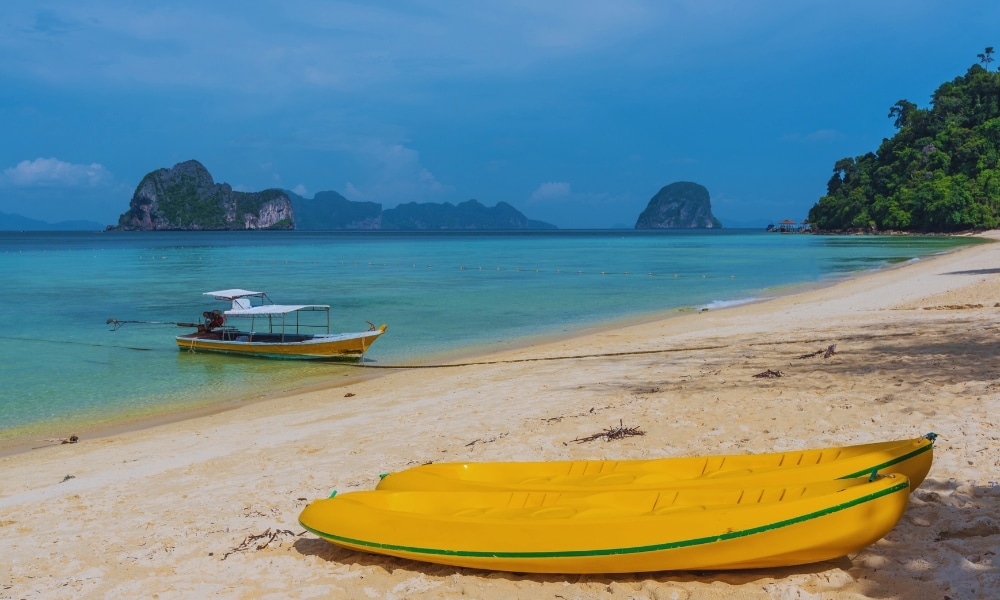 เรือคายัคสีเหลือง 2 ลำพักผ่อนบน ที่เที่ยวตรัง หาดทรายที่มีทะเลสีฟ้าครามอันเงียบสงบ ใกล้เรือในน้ำและเกาะห่างไกลภายใต้ท้องฟ้าแจ่มใส ถือเป็นภาพที่สมบูรณ์แบบของที่ท่องเที่ยวชุมพร.