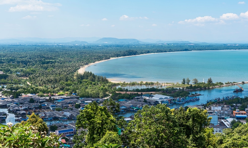 มุมมองทางอากาศของเมืองชายฝั่งชุมพรที่มีหาดทราย พืชพรรณอันเขียวชอุ่ม และ ท่องเที่ยวชุมพร อ่าวอันเงียบสงบ บ้านเรือนและเรือมองเห็นได้ใกล้ชายฝั่งภายใต้ท้องฟ้าสีครามสดใส ถือเป็นไฮไลท์ของที่ท่องเที่ยวชุมพร.