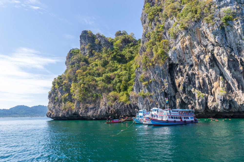 เรือที่ลอยอยู่ใกล้หน้าผาหินขนาดใหญ่ที่ปกคลุมไปด้วยแมกไม้เขียวขจีในวันที่อากาศสดใส มอบทิวทัศน์อัน ที่เที่ยวตรัง น่าหลงใหลชวนให้นึกถึงที่ตั้งท่องเที่ยวจังหวัดชุมพร ที่ซึ่งความงามของธรรมชาติมาบรรจบกับการผจญภัยอันไม่มีที่สิ้นสุด.