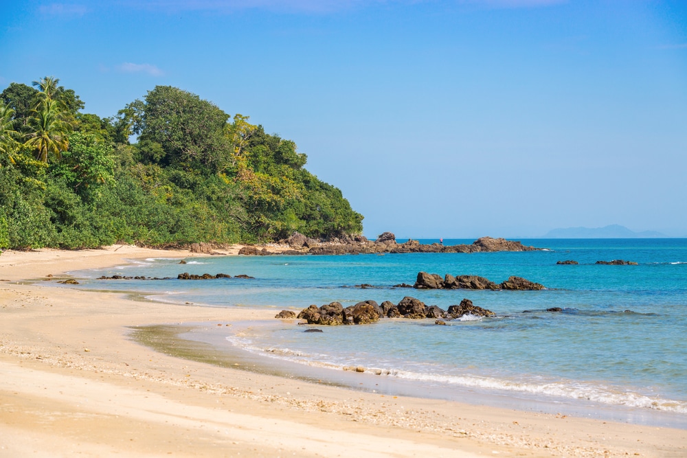 หาดทรายที่มีคลื่นอ่อนโยน แนวชายฝั่งหิน และต้นไม้เขียวชอุ่มใต้ท้องฟ้าสีฟ้า ที่เที่ยวตรัง ใส ทำให้เป็นสถานที่ท่องเที่ยวชุมพรในอุดมคติ.