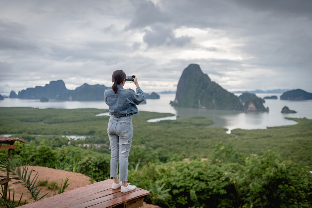 คนสวมชุดยีนส์ยืนอยู่บนแท่นไม้เต็นท์จุดกางพังงา กำลังถ่ายภาพทิวทัศน์ด้วยสมาร์ทโฟนที่เต็มไปด้วยต้นไม้เขียวขจีและเกาะหินใต้ท้องฟ้าที่มีเมฆครึ้ม.