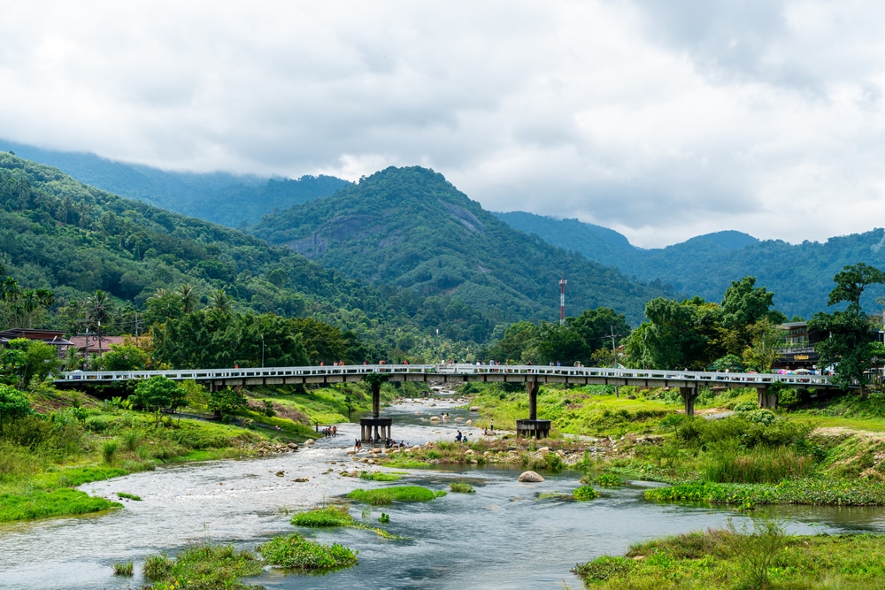 รถไฟข้ามสะพานข้ามแม่น้ำ โดยมีภูเขาสีเขียวชอุ่มและไร่กาแฟภาคเหนือเป็นพื้นหลังภายใต้ท้องฟ้าที่มีเมฆมาก. หมู่บ้านสวยในไทย 