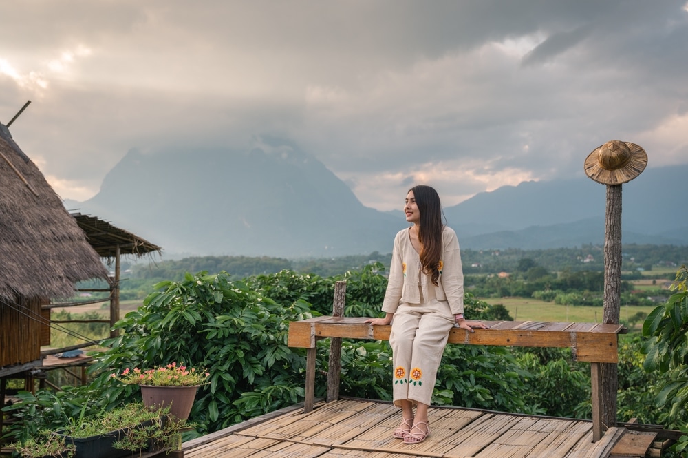 สตรีคนหนึ่งนั่ง หมู่บ้านสวยในไทย อยู่บนม้านั่งไม้ รายล้อมไปด้วยความงามอันเงียบสงบของไร่กาแฟภาคเหนือ พร้อมด้วยขุนเขาอันยิ่งใหญ่และท้องฟ้าที่มีเมฆปกคลุมเป็นฉากหลัง.