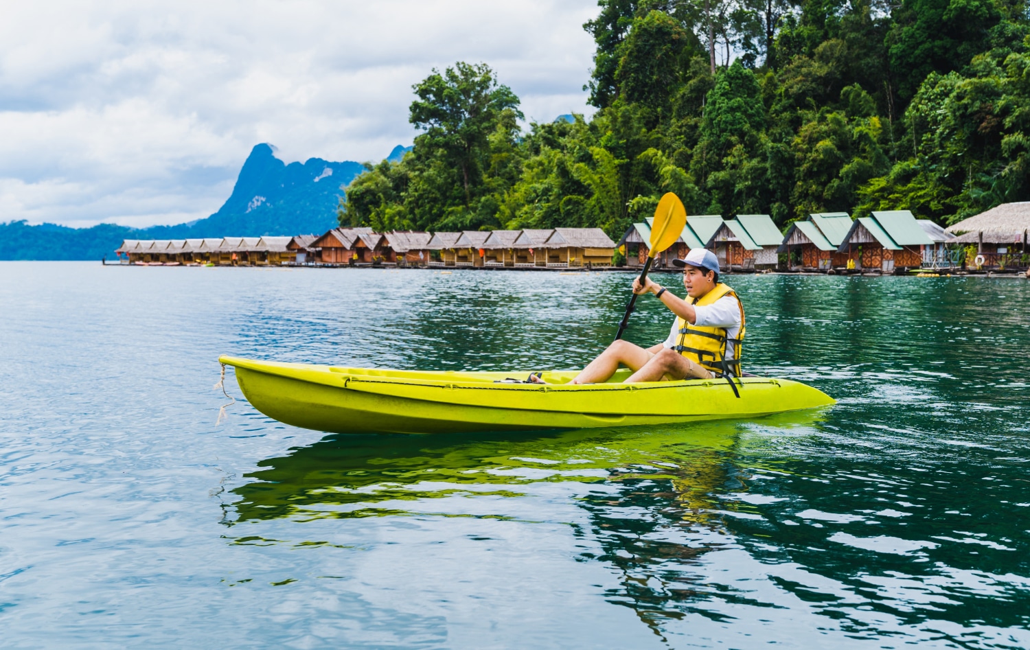 คนหนึ่งกำลังพายเรือคายัคสีเหลืองในทะเลสาบที่มีบ้านลอยน้ำและเนินเขาที่เป็นป่าเป็นฉากหลัง จับภาพความงามอันเงียบสงบของที่เที่ยวปลายฝนต้น ที่เที่ยวปลายฝนต้นหนาว หนาว.