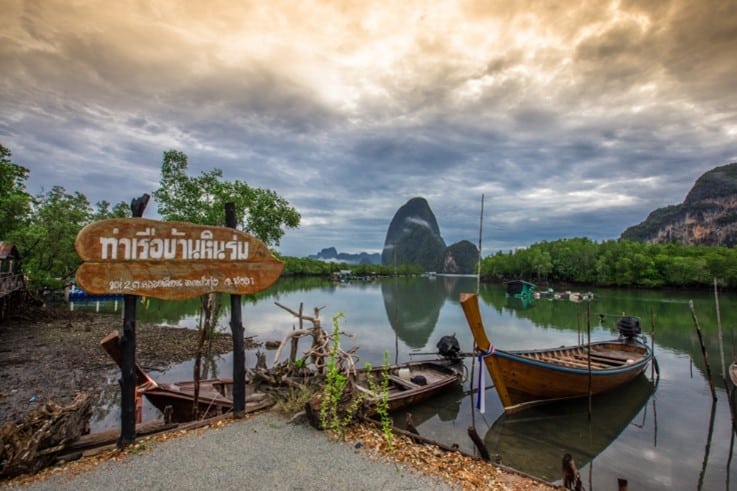 วิวแม่น้ำอันเงียบสงบที่มีเรือจอดอยู่ท่ามกลางแมกไม้เขียวขจี มีภูเขาเป็นฉากหลังชวนให้นึกถึง หมู่บ้านสวยในไทย และป้ายไม้บนชายฝั่ง.