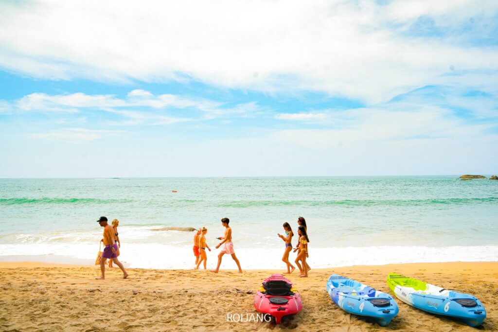 ผู้คนเดินเล่นไปตามหาดทรายใกล้ทะเล โดยมีเรือคายัคสีน้ำเงินและสีแดงจอดพักอยู่ริมฝั่งใต้ท้องฟ้าที่มีเมฆบางส่วน ใกล้ๆ กันนั้น กลิ่นหอมชวนเชิญจากครัวบางสักรับประกันว่าคุณจะได้ลิ้มรสอาหารท้องถิ่นรสเลิศ.