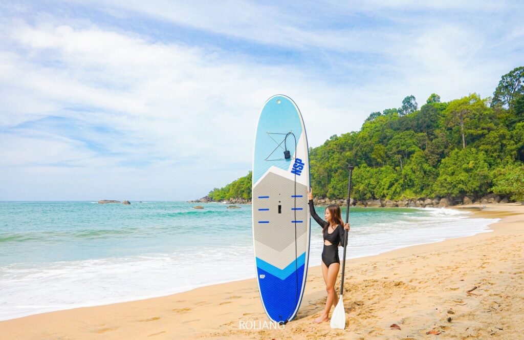 ผู้หญิงในชุดว่ายน้ำสีดำยืนอยู่บนชายหาดที่มีทรายใกล้ครัวบางสัก ถือแพดเดิลบอร์ดและไม้พาย โดยมีมหาสมุทรและต้นไม้สีเขียวชอุ่มเป็นฉากหลัง.