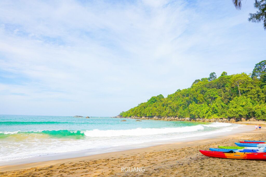 ชายหาดที่เงียบสงบมีคลื่นสีฟ้าคราม ชายหาดทราย และเนินเขาเขียวขจีเป็นฉากหลัง มีเรือคายัคเรียงรายอยู่บนพื้นทรายใกล้กับครัวบางสัก ซึ่งเป็นจุดที่สมบูรณ์แบบสำหรับการผจญภัยและการพักผ่อนท่ามกลางความงามตามธรรมชาติอันเงียบสงบ.