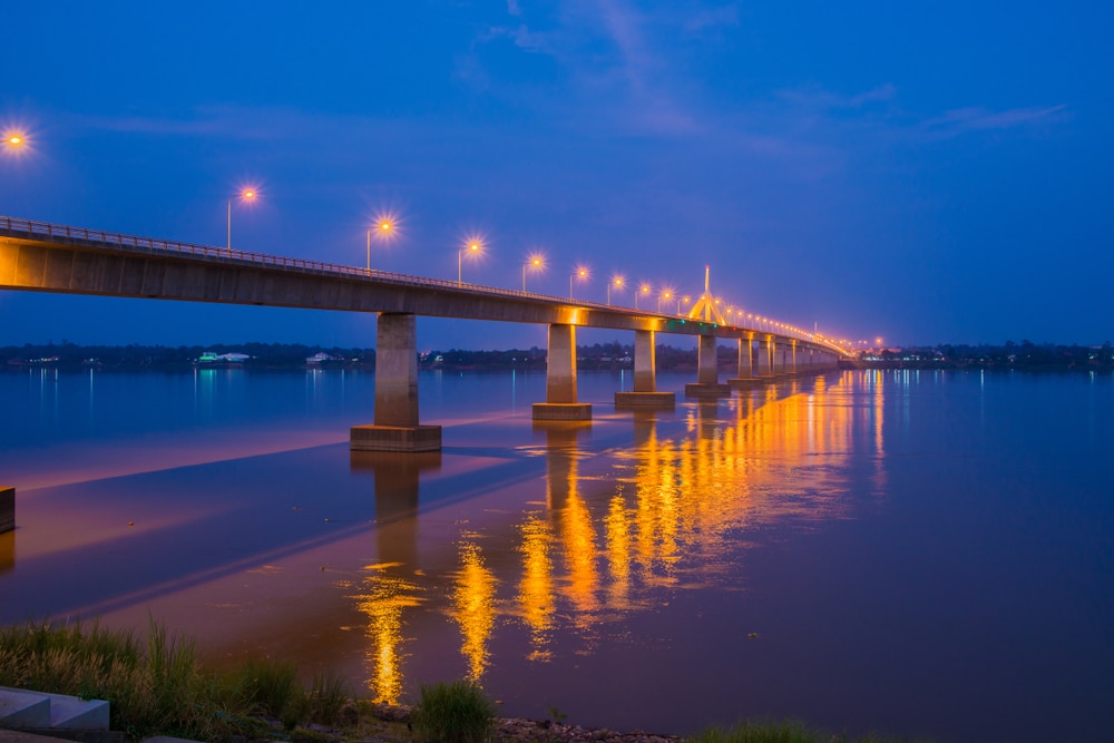 สะพานยาวที่มีแสงไฟถนนส่องสว่างสะท้อนลงในแม่น้ำอันเงียบสงบภาย วัดภูมโนรมย์ ใต้ท้องฟ้าพลบค่ำ สร้างทัศนียภาพอันเงียบสงบที่ชวนให้นึกถึงความเงียบสงบที่พบใกล้กับวัดภูมโนรมย์.