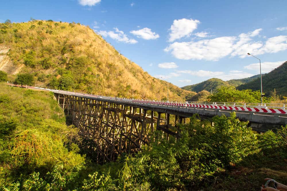 สะพานที่มีราวกั้นทอดข้ามหุบเขาสี ที่เที่ยวเพชรบูรณ์ เขียวขจี มองเห็นทิวเขาที่ปกคลุมไปด้วยพืชพรรณแห้งภายใต้ท้องฟ้าสีฟ้าสดใส จุดชมวิวอันเงียบสงบแห่งนี้เป็นตัวอย่างความงามตามธรรมชาติที่พบได้ใน ที่เที่ยวระนอง.