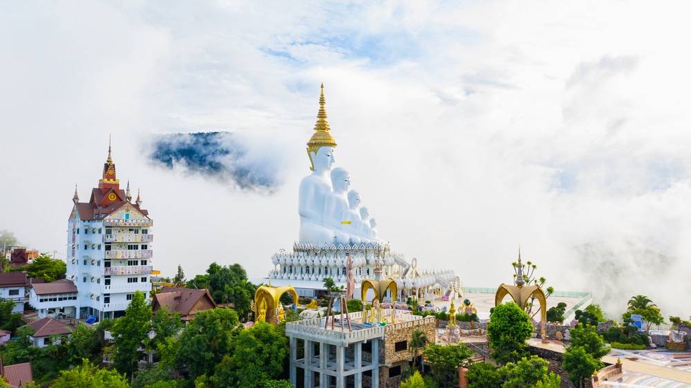 มุมมองทางอากาศของวัดอันเงียบ ที่เที่ยวเพชรบูรณ์ สงบที่มีพระพุทธรูปหลายองค์เป็นสีขาว โอบล้อมไปด้วยแมกไม้เขียวขจีและหมอก อาคารแบบดั้งเดิมตั้งอยู่ทางด้านซ้าย มองเห็นสถานที่ท่องเที่ยวระนองอันน่าหลงใหล.