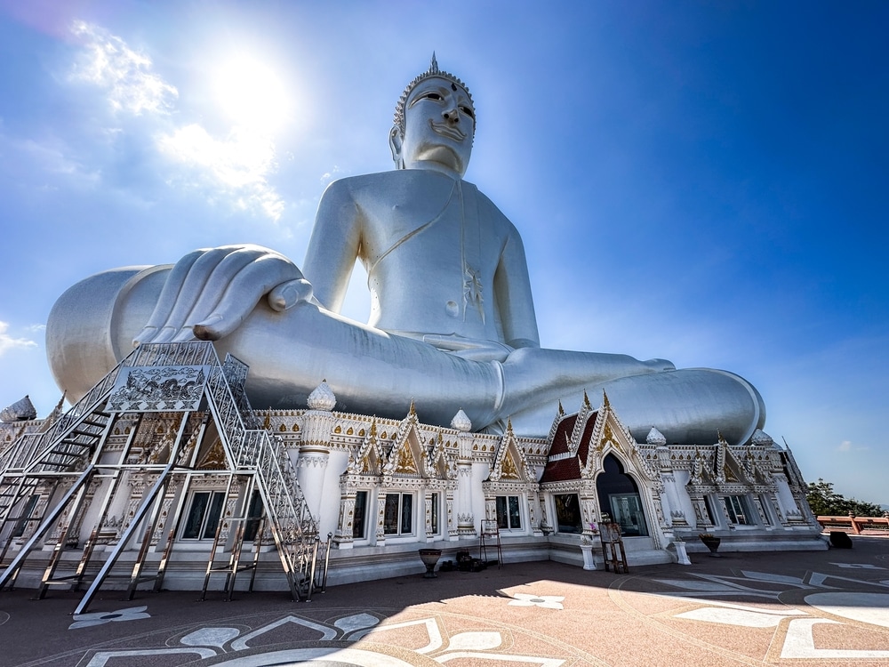 พระพุทธรูปสีขาวคู่บารมีของวัดภูมโนรมย์นั่งอย่างเงียบสงบภายใต้ท้องฟ้าสีฟ้าใส ฐานที่วิจิตรวิจิตรและบันไดที่เชิญชวนผู้มาเยือนขึ้นไป. ภูมโนรมย์