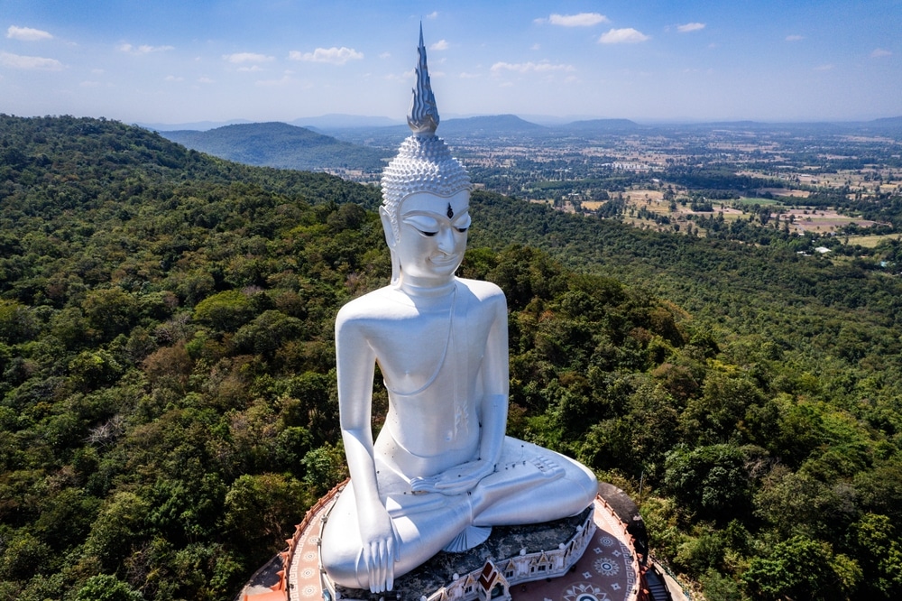 มุมมองทางอากาศของพระพุทธรูปสี วัดภูมโนรมย์ ขาวองค์ใหญ่ประทับนั่งสมาธิอยู่บนยอดเขาภูมโนรมย์อันเงียบสงบ รายล้อมไปด้วยต้นไม้เขียวขจี และมองเห็นทิวทัศน์อันกว้างใหญ่ภายใต้ท้องฟ้าสีฟ้าที่มีเมฆบางส่วน.