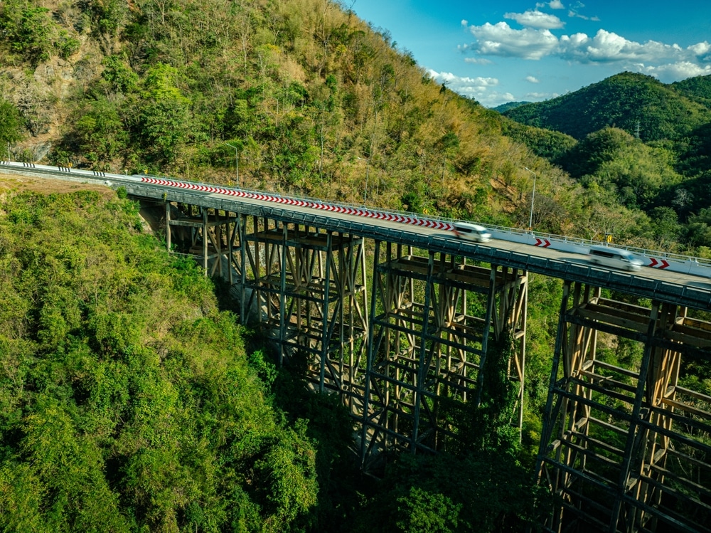 สะพานยาวทอดข้ามภูมิประเทศที่ ที่เที่ยวเพชรบูรณ์ เขียวขจีบนเนินเขาในเขตจังหวัดระนองที่งดงาม สะพานนี้มีโครงเหล็กรองรับใต้สะพาน และมองเห็นรถยนต์ไม่กี่คันบนถนนด้านบน ทำให้ผู้เดินทางมองเห็นเมืองระนองในมุมมองที่ไม่เหมือนใคร.