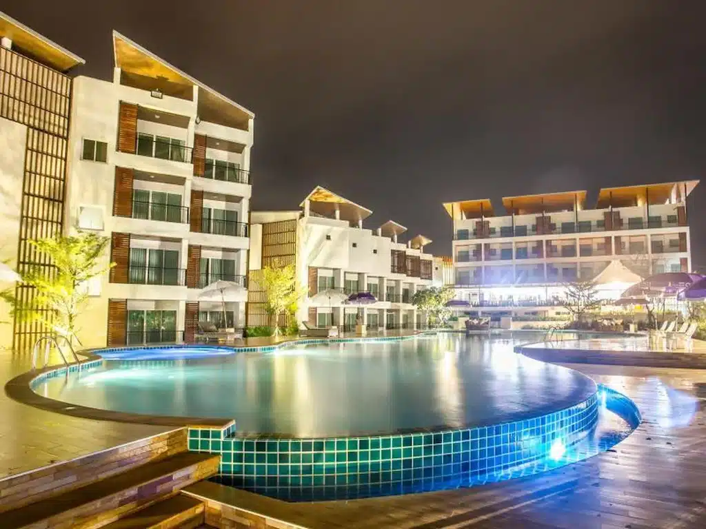 ทิวทัศน์ยามค่ำคืนแสดงให้เห็นโรงแรมทันสมัยที่หาดเจ้าหลาวติด ที่พักหาดเจ้าหลาว ทะเล โดยมีระเบียงที่ส่องสว่างซึ่งสะท้อนจากสระว่ายน้ำทรงกลมขนาดใหญ่ที่อยู่เบื้องหน้า สร้างบรรยากาศริมชายหาดอันเงียบสงบ.