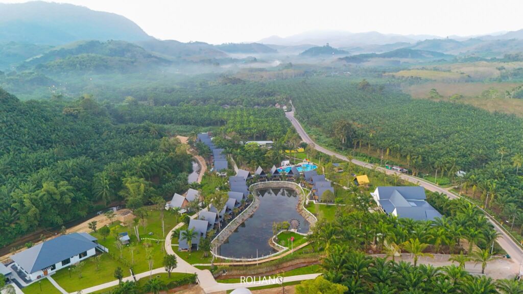 ภาพมุมสูงของภูมิประเทศชนบทเผยให้เห็นกลุ่มอาคารที่ซ่อนตัวอยู่รอบสระน้ำ ล้อมรอบด้วยต้นไม้เขียวขจีและเนินเขาไกลๆ ภายใต้ท้องฟ้าที่มีหมอก ชวนให้นึกถึงเสน่ห์อันเงียบสงบของผืนน้ำอันเงียบสงบของ เช่าเรือเขาหลัก.