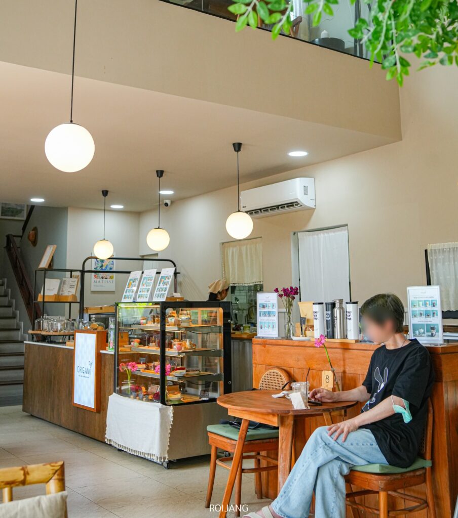 มีคนนั่งอยู่ที่โต๊ะไม้ใน Origami Cafe Bangtao ซึ่งเป็นร้านบรรยากาศสบายๆ ที่มีคาเฟ่น่ารักย่านบางเทา ภูเก็ต ล้อมรอบด้วยแผงขายขนมอันน่ารื่นรมย์และบอร์ดเมนูเป็นฉากหลัง.