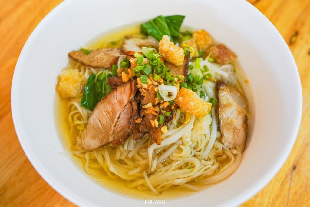 ก๋วยเตี๋ยวน้ำใสชามโตที่มีหมูแผ่น ผักใบเขียว ถั่วงอก กระเทียมเจียว ต้นหอมสับ และหนังหมูกรอบวางอยู่บนโต๊ะไม้อย่างน่าทานที่บะหมี่พังงาตลาดขวาง.