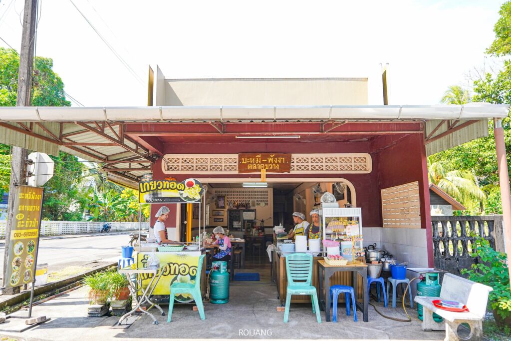 ร้านขายอาหารริมทางในบะหมี่พังงาตลาดขวางเปิดแผงขายอาหารในร่มบรรยากาศอบอุ่นพร้อมเสิร์ฟอาหารหลากหลายชนิด รอบๆ แผงขายอาหารมีเก้าอี้และโต๊ะ คนหนึ่งนั่งดูแลเคาน์เตอร์ ขณะที่อีกคนเตรียมอาหาร โดยมีฉากหลังเป็นต้นไม้ใบเขียวขจี.