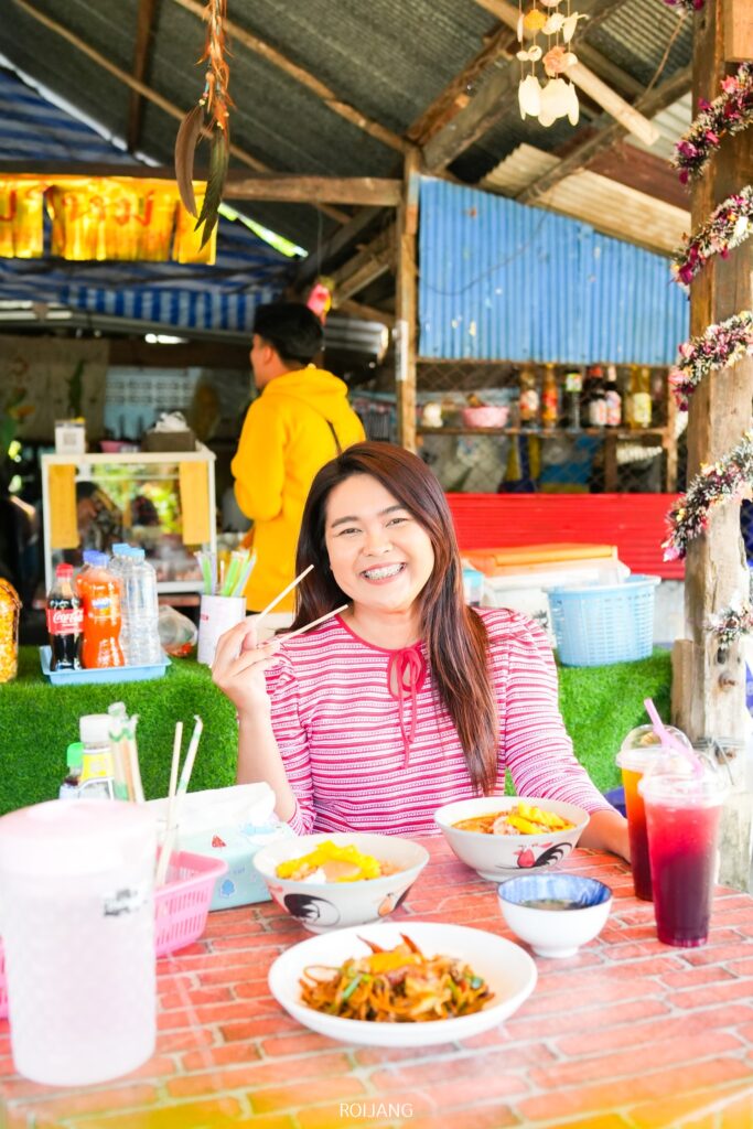 หญิงสาวสวมเสื้อเชิ้ตลายทางสีแดงยิ้มขณะเพลิดเพลินกับก๋วยเตี๋ยวและเครื่องดื่มที่โต๊ะกลางแจ้ง ชวนให้นึกถึงช่วงเย็นที่คึกคักที่ นับดาวเย็นตาโฟ.