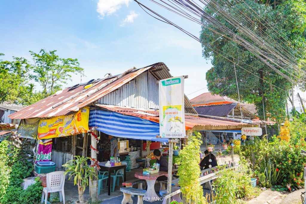 ร้านอาหารกลางแจ้งขนาดเล็ก นับดาวเย็นตาโฟ มีหลังคาโลหะและผ้าใบสีสันสดใส ล้อมรอบด้วยต้นไม้เขียวชอุ่ม ผู้คนต่างเพลิดเพลินกับมื้ออาหารที่โต๊ะ ขณะที่สายไฟเหนือศีรษะทอดยาวไขว้กันใต้ต้นไม้ในพื้นหลัง.